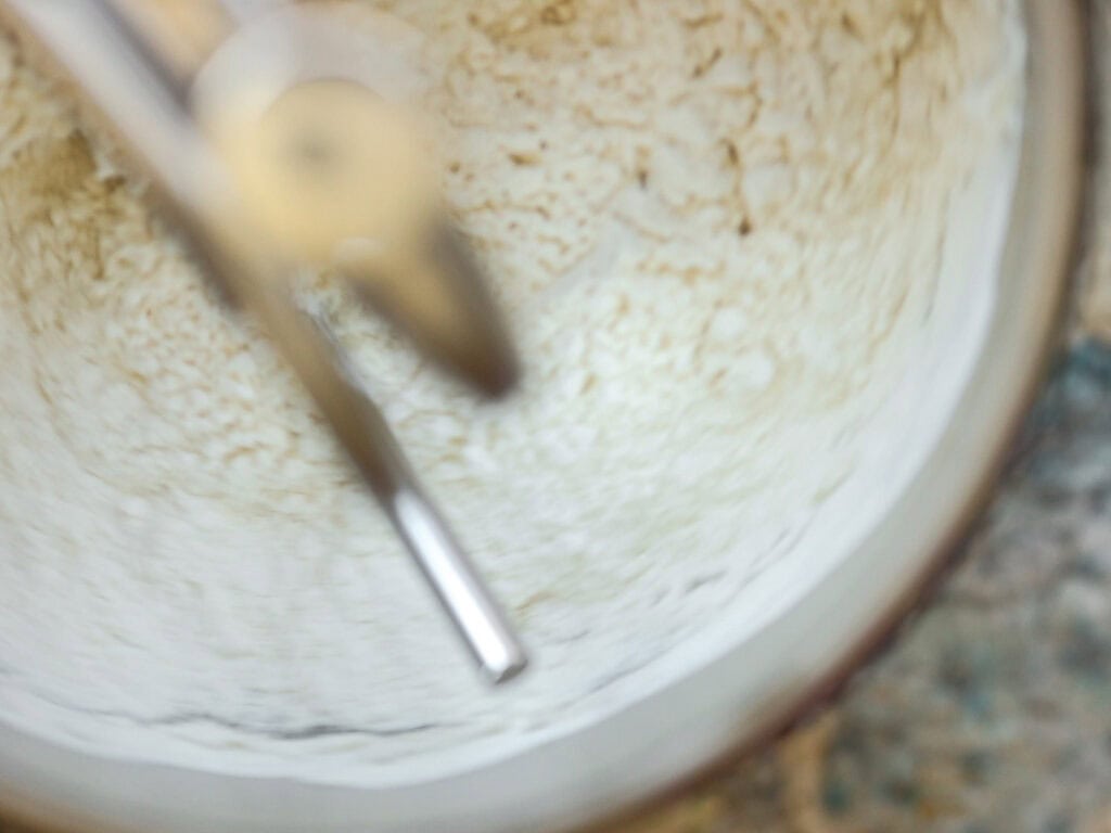 A close-up of a mixer in a mixing bowl with a dough being stirred. The motion blur indicates the mixer is in use, blending the ingredients. The bowl is partially visible against a speckled countertop.